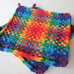 May include: A rainbow colored crocheted pot holder with a woven pattern and a purple border.