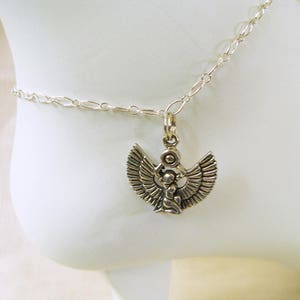 May include: Silver-toned anklet featuring a detailed pendant of a winged figure, possibly an Egyptian deity. The pendant is suspended from a delicate chain. The anklet is shown on an ankle, highlighting the jewelry's design and placement.