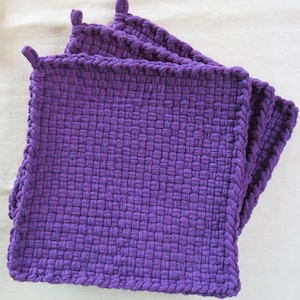May include: A stack of three square, woven potholders in a vibrant purple hue. Each potholder features a looped corner for hanging and a tightly woven texture. The potholders are made of a thick, durable material.