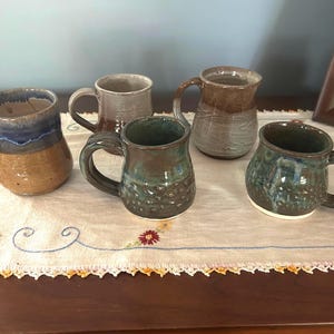 May include: A collection of five ceramic mugs with various glaze finishes, including blue, brown, grey, and green tones. Each mug features a rounded body, a sturdy handle, and a unique textured surface, displayed together as a set of drinkware.