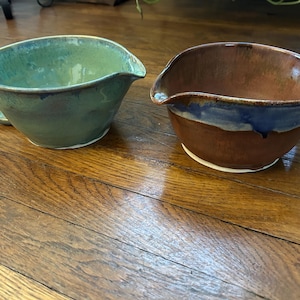 Può includere: Due ciotole in ceramica per mescolare con beccucci. Una è verde con manico, l'altra è marrone con bordo blu. Entrambe le ciotole poggiano su una base bianca e su una superficie di legno.