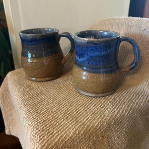May include: Two handmade ceramic mugs. The mugs have a brown base and a blue top, with a glossy finish. Each mug has a curved handle. The mugs are sitting on a tan towel.