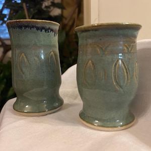 Puede incluir: Dos tazas de cerámica verde con un diseño texturizado. Las tazas tienen un esmalte verde claro con un borde verde oscuro y un acento marrón alrededor de la parte superior.