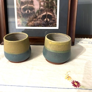 May include: Two handmade ceramic cups. Each cup has a rounded shape with a base of reddish-brown, a band of blue-green in the middle, and a yellow-tan top. The cups are displayed on a white tablecloth with floral embroidery, with a framed picture of raccoons in the background.