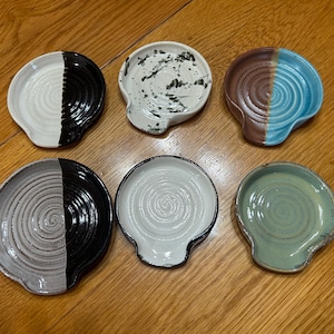 May include: Six ceramic spoon rests in assorted colours and patterns. Each rest has a circular design with a small handle. The colour schemes include black and white, brown and blue, and speckled patterns. The rests are displayed on a wooden surface.