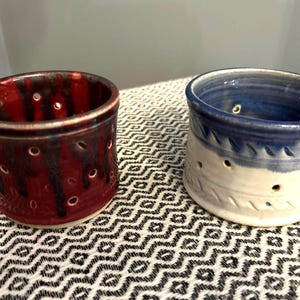 May include: Two handmade ceramic candle holders. One is a deep red with dark streaks and the other is blue and white. Both have a cylindrical shape with holes and decorative patterns.