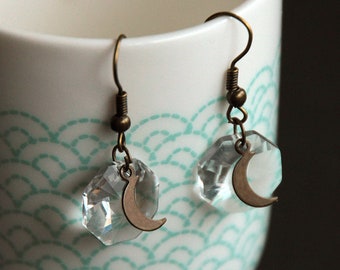 May include: A pair of dangle earrings with clear crystal teardrop-shaped charms and crescent moon accents. The earrings are made with bronze-colored metal.