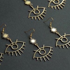 May include: Four pairs of gold-tone earrings with a dangling eye design and a starburst charm. The earrings are made of metal and have a delicate, feminine look.