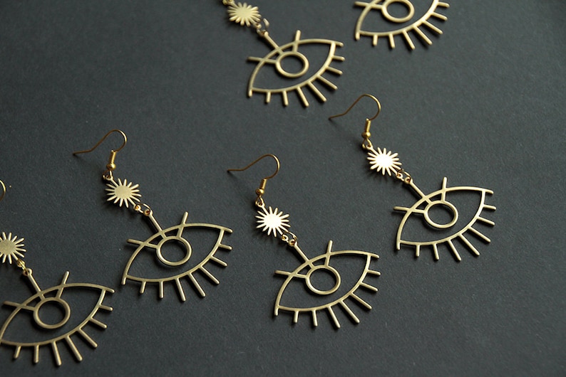 May include: Four pairs of gold-tone earrings with a dangling eye design and a starburst charm. The earrings are made of metal and have a delicate, feminine look.