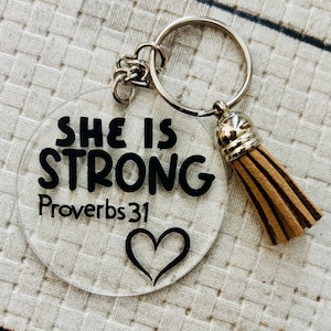 以下が含まれることがあります： 黒い文字で「She is Strong Proverbs 31」と書いてあり、その下に黒いハートが描かれた透明な丸いキーホルダー。キーホルダーには銀色の金属製のリングと茶色のタッセルが付いています。