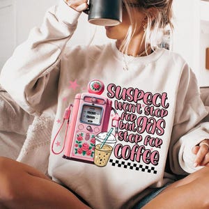 Puede incluir: Sudadera blanca con una bomba de gasolina rosa y una taza de café. El texto en la sudadera dice "Suspect won't stop for gas but stop for coffee".