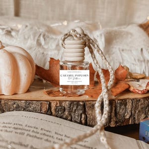 Op de afbeelding: Een kleine, transparante glazen fles met een houten dop, met het opschrift "CARAMEL POPCORN", hangt aan een touw. Het staat op een houten schijf met herfstbladeren en een pompoen. Een open boek ligt op de voorgrond.