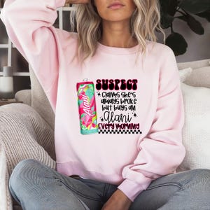 Puede incluir: Sudadera rosa con un gráfico de una lata de bebida energética Alani Nu y el texto "SUSPECT Claims she's always broke but buys an Alani every morning".