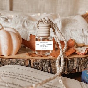 May include: A clear glass bottle with a wooden cap and a white label that reads "SWEATER WEATHER". The bottle is on a wooden surface with fall leaves and a decorative pumpkin. A book is open in the foreground.