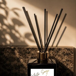 Può includere: Un diffusore quadrato nero con un collare color oro, riempito con bastoncini neri. L'etichetta recita "White Sage & Lavender" in una scrittura elegante. Il prodotto è un diffusore a lamelle di lusso.