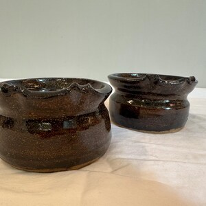 May include: Two dark brown ceramic bowls with a textured, speckled glaze. The bowls have a slightly rounded, oval shape and a wavy rim.