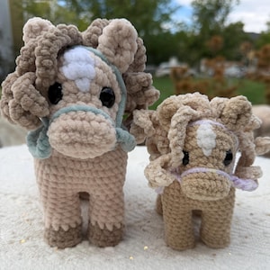 May include: Two crocheted plush horses made from tan yarn with curly manes, white forehead patches, and black plastic eyes. The larger horse wears a light blue halter, while the smaller horse wears a light purple halter, both featuring soft, textured stitches.