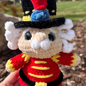 May include: A crocheted nutcracker doll featuring a black hat with a red plume and blue accent, white curly hair, a beige face with large dark eyes, and a red jacket with yellow horizontal stripes and a black belt with a yellow buckle.