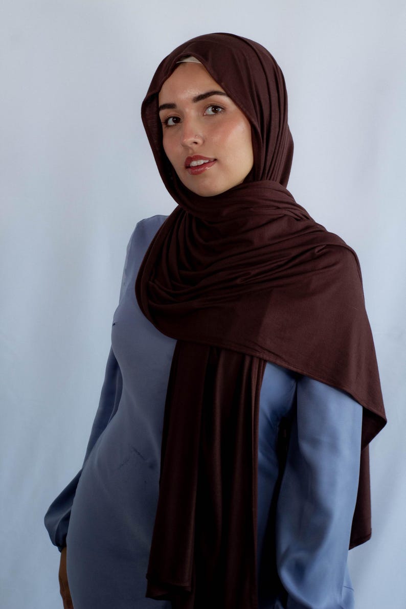 K&ouml;nnte beinhalten: Eine Frau tr&auml;gt ein braunes Jersey-Hijab. Das Hijab ist &uuml;ber Kopf und Schultern gelegt, und sie tr&auml;gt ein hellblaues Hemd.