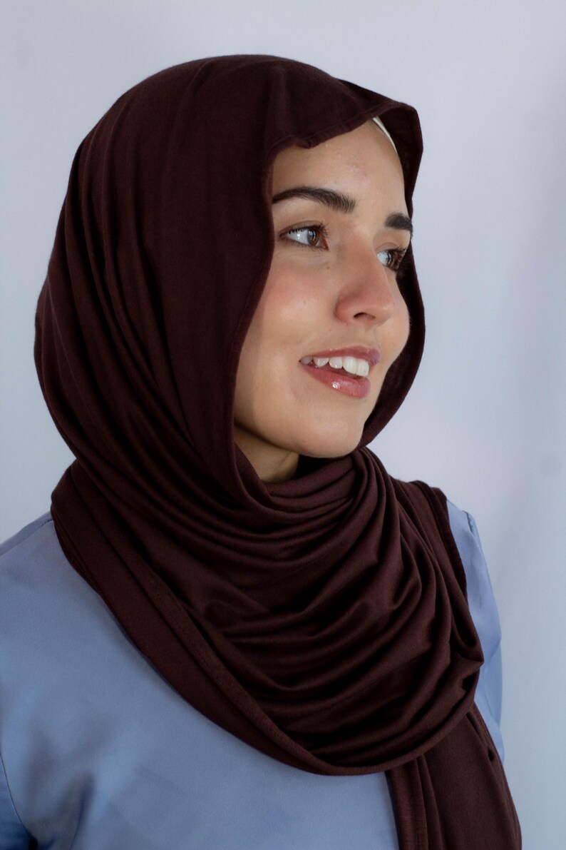 K&ouml;nnte beinhalten: Ein braunes Jersey-Hijab, getragen von einer Person mit braunen Augen und einem hellblauen Hemd.