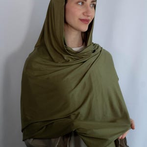 K&ouml;nnte beinhalten: Ein langer, olivgr&uuml;ner Kapuzen-Schal aus weichem Stoff. Der Schal ist um den Hals und die Schultern einer Person gelegt, mit der Kapuze nach oben gezogen.