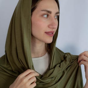 K&ouml;nnte beinhalten: Eine Person tr&auml;gt einen gr&uuml;nen Kapuzen-Schal. Der Schal ist aus weichem, dehnbarem Stoff und kann auf verschiedene Arten getragen werden.