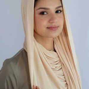 K&ouml;nnte beinhalten: Ein cremefarbener Jersey-Hijab mit Kapuze. Der Hijab ist um Hals und Schultern gelegt, die Kapuze ist &uuml;ber den Kopf gezogen.