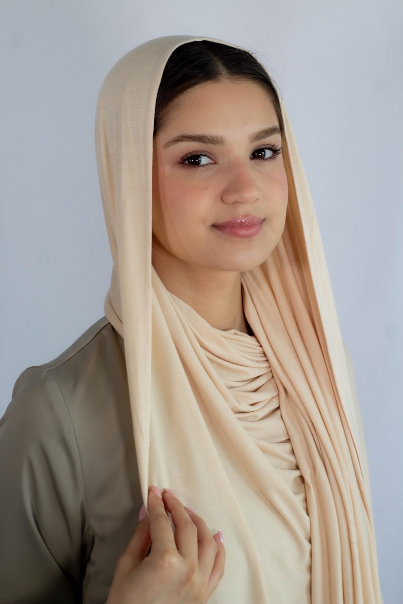 K&ouml;nnte beinhalten: Ein cremefarbener Jersey-Hijab mit Kapuze. Der Hijab ist um Hals und Schultern gelegt, die Kapuze ist &uuml;ber den Kopf gezogen.