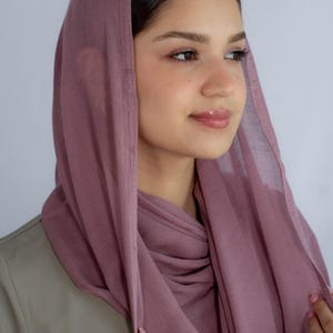 May include: A woman wearing a mauve-colored headscarf. The scarf is draped over her head and shoulders, and she is holding it in place with her hands.
