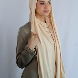 K&ouml;nnte beinhalten: Eine Frau tr&auml;gt einen hellbeigen Jersey-Hijab. Der Hijab ist &uuml;ber Kopf und Hals gelegt, und der Stoff ist vorne zusammengezogen. Die Frau tr&auml;gt ein lang&auml;rmeliges, hellbraunes Hemd.