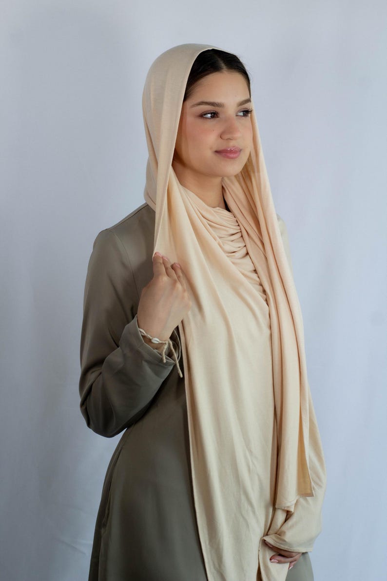 K&ouml;nnte beinhalten: Eine Frau tr&auml;gt einen hellbeigen Jersey-Hijab. Der Hijab ist &uuml;ber Kopf und Hals gelegt, und der Stoff ist vorne zusammengezogen. Die Frau tr&auml;gt ein lang&auml;rmeliges, hellbraunes Hemd.