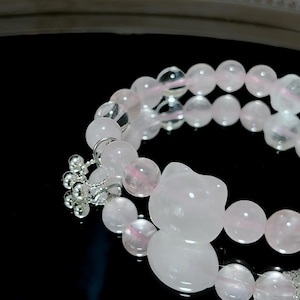 Könnte beinhalten: Ein Armband aus Rosenquarzperlen mit einem Silberanhänger und einem Katzenanhänger aus Rosenquarz. Das Armband hat runde Perlen und einen katzenförmigen Anhänger. Der Text "Embedded Studio" befindet sich am unteren Bildrand.