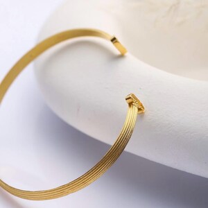 Puede incluir: Una pulsera de puño abierta de color dorado con un diseño texturizado y acanalado. La pulsera es curva y tiene un estilo simple y elegante. La joya se muestra sobre un fondo blanco.