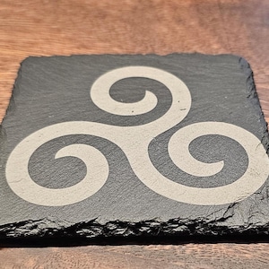 May include: A black slate coaster with a white Celtic knot design. The coaster has a rough, natural edge.