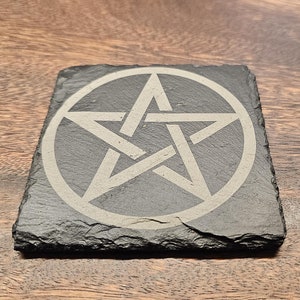 Pode incluir: Uma base de ardósia cinza escuro quadrada com um design de pentagrama cinza claro. O pentagrama está dentro de um círculo. A base tem uma superfície áspera e texturizada, sobre uma superfície de madeira.