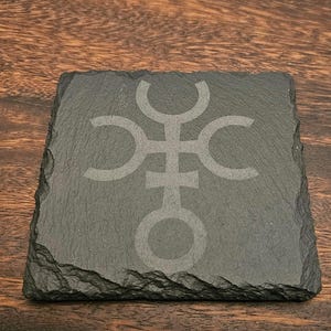 Könnte beinhalten: Ein schwarzer Schieferuntersetzer mit einem weißen Symbol eines Kreises mit einem Kreuz und drei Halbmonden.