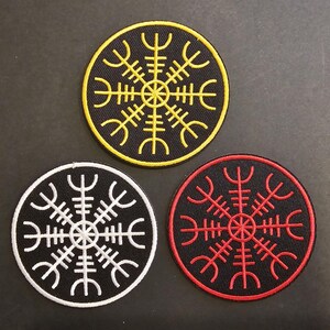 Peut inclure: Trois patchs brodés avec un fond noir et un symbole blanc, jaune ou rouge. Le symbole est une étoile stylisée à huit branches avec un cercle central.