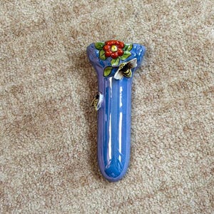 May include: A blue ceramic vase with a flower and bee design. The vase has a long, slender shape with a flared top. A red flower with green leaves and two bees are on the vase. The vase is approximately 8 inches tall.