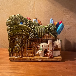 May include: A brown ceramic pen holder with the words "Under the Spreading Chestnut Tree." The holder features a detailed relief scene with figures, a horse, and a tree. Pens and markers are visible inside the holder.