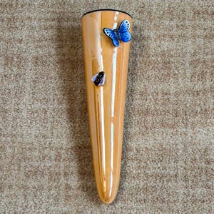 May include: A tall, orange-toned vase with a glossy finish. The vase is adorned with a blue butterfly and a bee. The vase has a black rim and a tapered shape, suitable for displaying flowers or decorative branches.