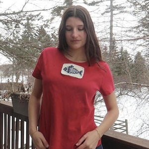 Op de afbeelding: Een rood T-shirt met korte mouwen en een rechthoekige patch met een blauw en wit visontwerp. De patch bevindt zich op de borst. De achtergrond toont een houten terras en bomen.