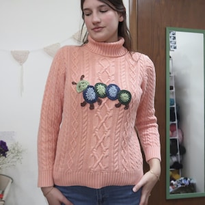 May include: A peach-colored cable-knit turtleneck sweater with a crocheted caterpillar design on the front. The caterpillar features green, blue, and brown accents. The sweater has long sleeves and a ribbed collar.