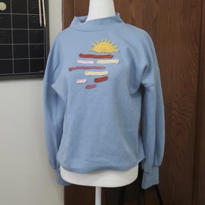 Op de afbeelding: Een lichtblauw sweatshirt met een hoge kraag en lange mouwen. De voorkant heeft een gehaakt geel zonontwerp boven horizontale lijnen in verschillende kleuren. Het sweatshirt wordt getoond op een witte mannequin.
