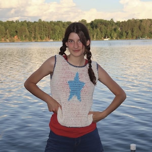 May include: A cream-colored, sleeveless crochet top with a large blue star design. The top has a blue trim and is worn over a red shirt. The person is standing in front of a lake.