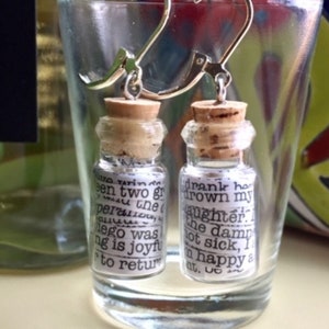 May include: A pair of silver earrings with small glass bottles filled with paper scraps. The bottles are corked and have text printed on the paper scraps. The text reads "drank ho drown my daughter the damn not sick, I am happy &" and "een two gr per the lego was ng is joyft to return".