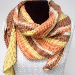 May include: A striped knitted scarf in shades of peach, yellow, brown, and white. The scarf is folded and draped over a white mannequin.