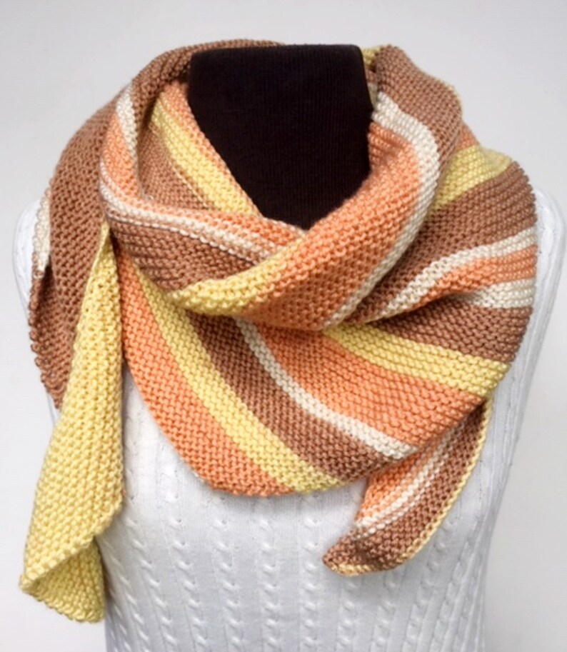 May include: A striped knitted scarf in shades of peach, yellow, brown, and white. The scarf is folded and draped over a white mannequin.