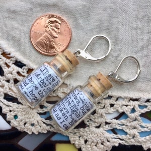 May include: A pair of silver earring hooks with small glass bottles attached. The bottles have cork stoppers and contain handwritten messages. The message on the left bottle reads "There is a little &iexcl;&Aacute;rbol de la vida! I hope the..." and the message on the right bottle reads "...stay ante rows, but I have strengthl from..."