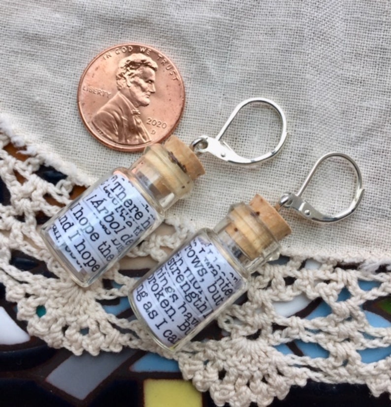 May include: A pair of silver earring hooks with small glass bottles attached. The bottles have cork stoppers and contain handwritten messages. The message on the left bottle reads "There is a little &iexcl;&Aacute;rbol de la vida! I hope the..." and the message on the right bottle reads "...stay ante rows, but I have strengthl from..."