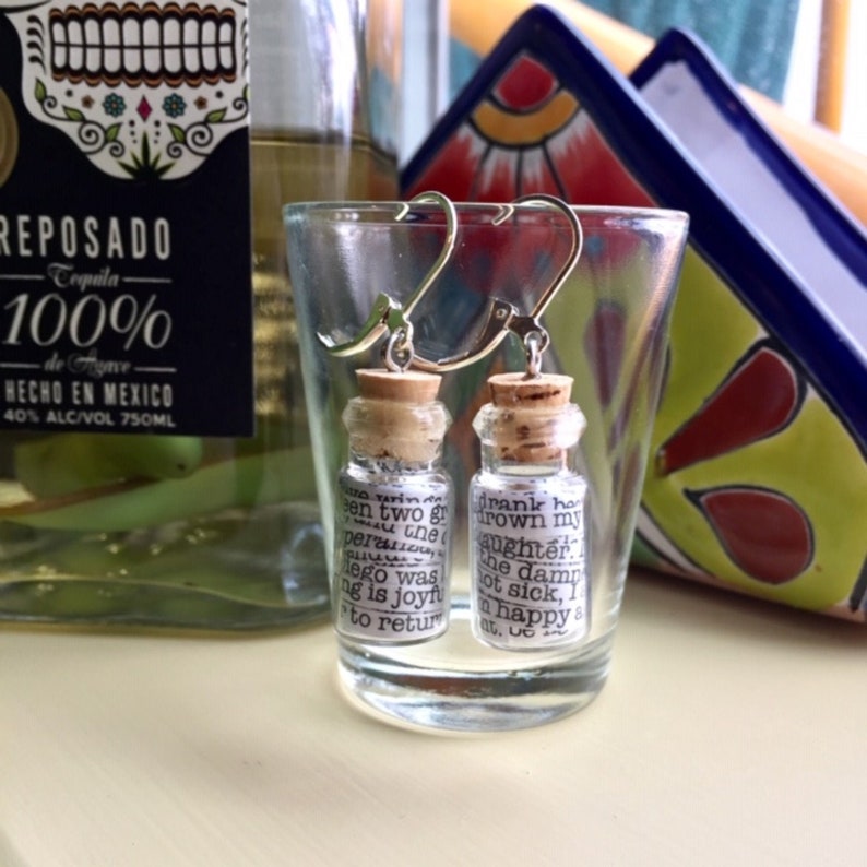 May include: A pair of silver earrings with small glass bottles filled with paper scraps. The bottles are corked and have text printed on the paper scraps. The text reads "drank ho drown my daughter the damn not sick, I am happy &" and "een two gr per the lego was ng is joyft to return".